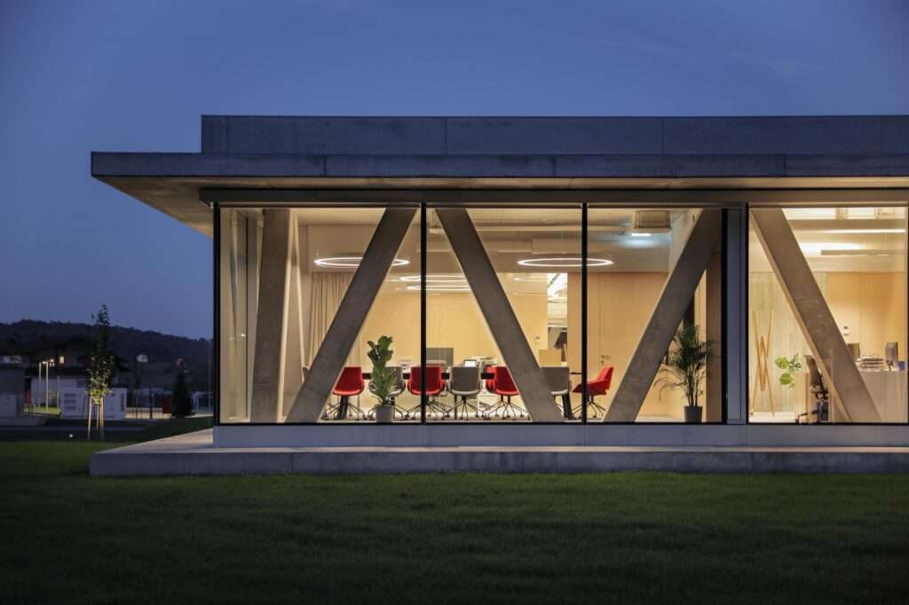 Dusk view of the lit meeting room inside the Pina Astra office showing V-shaped structural concrete columns through transparent glass windows.