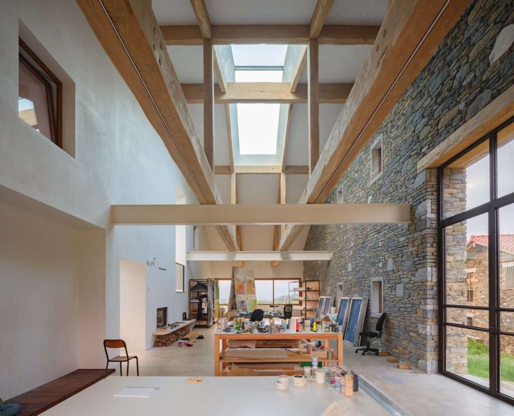 Double height artist studio and workshop inside Casa Basque House with an original stone wall, modern white partitions, exposed timber beams, and a central skylight.