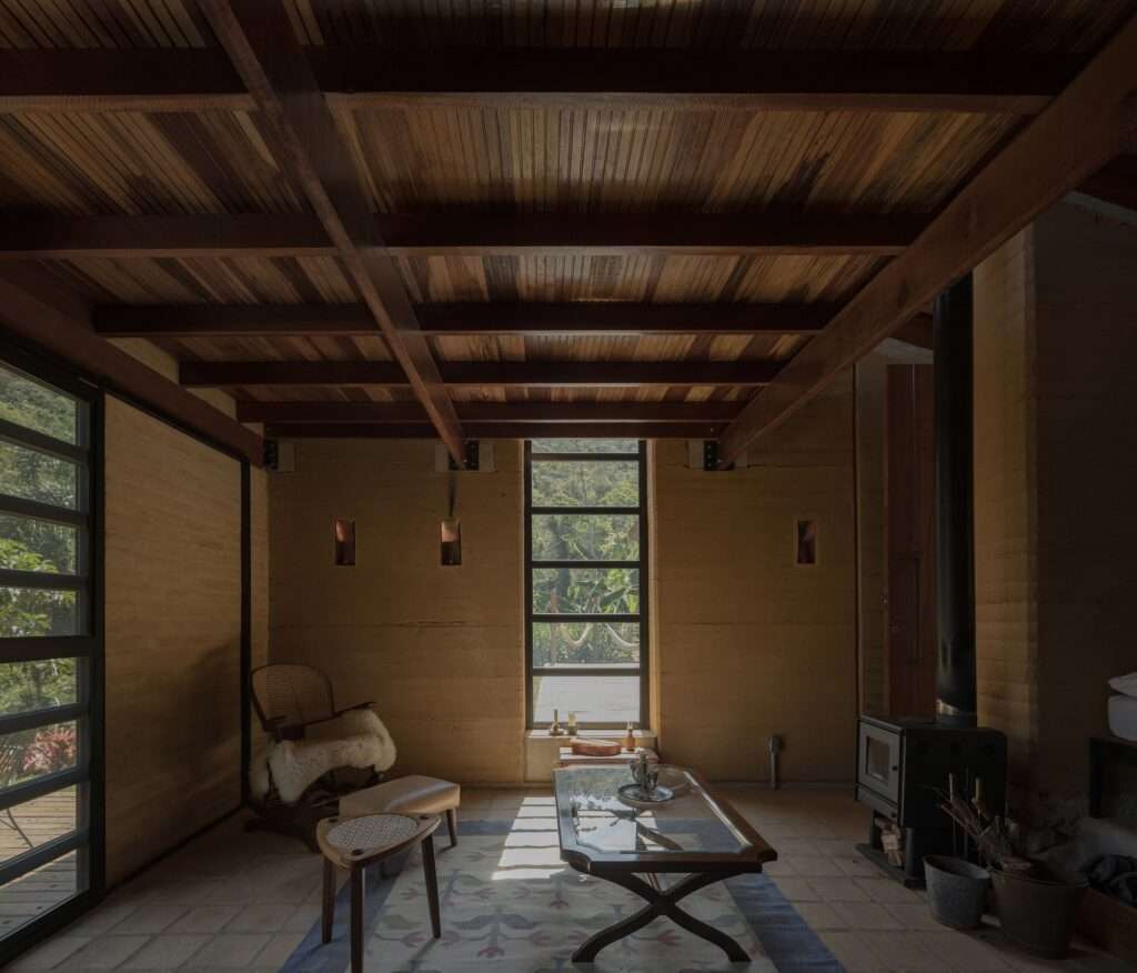 Interior view of the living area, featuring warm, textured rammed earth walls, a wooden ceiling, and floor-to-ceiling glass doors overlooking the forest.