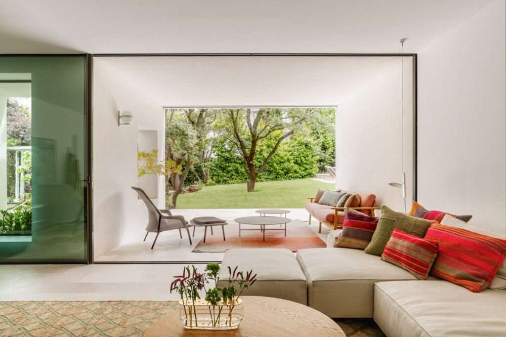 An interior view of a modern living room with a neutral-colored L-shaped sofa and colorful pillows, a gray lounge chair, and a low coffee table, opening entirely to the garden via massive sliding glass doors.