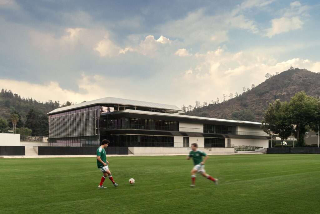 Soccer players training on a lush green pitch with the modern multi-tiered athletic pavilion and a mountainous landscape in the background.