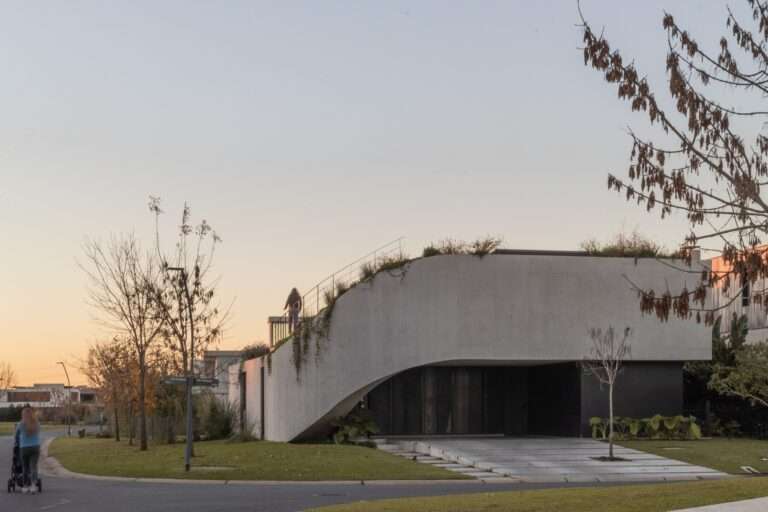 A brutalist-inspired residential facade featuring a sweeping curved concrete wall, integrated landscape planters, and an external volumetric staircase leading to a green roof.