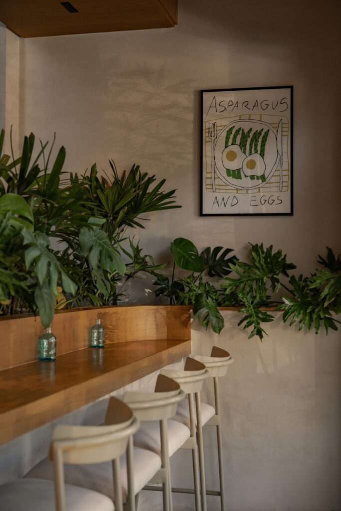 High-top wooden counter seating with barstools nestled against lush indoor planters and framed artwork titled Asparagus and Eggs.