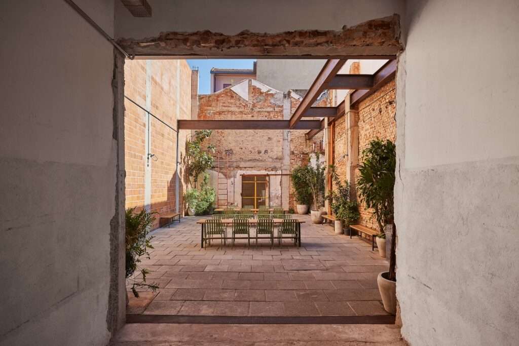 Fábrica Bhering adaptive reuse in Rio de Janeiro, Brazil. Perspective view from inside looking out through a large opening into the outdoor courtyard with a long communal table, exposed brick walls, and industrial steel beams. Potted plants line the walls. Photographed by Rafael Salim.