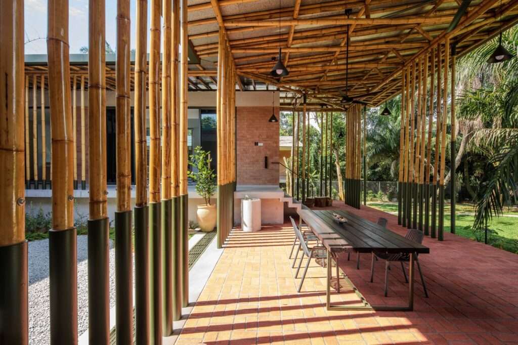 Shaded semi-outdoor verandah at Anjung House featuring rhythmic bamboo columns, brick flooring, a long wooden dining table, and dynamic daylight filtering through.