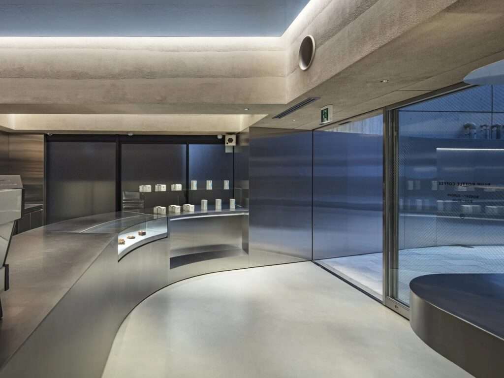 Curved stainless steel service counter inside Blue Bottle Coffee Harajuku by TEKI Design with indirect lighting and glass entrance.