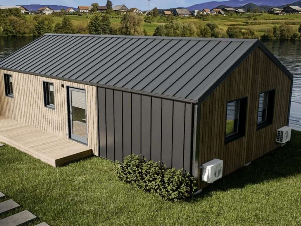 An elevated exterior view of the Vagabond Haven Evergreen XL prefab home, showcasing its natural wood cladding, dark metal standing seam roof, and large glass windows, set on green grass near a lake with a village in the background.