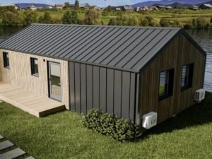 An elevated exterior view of the Vagabond Haven Evergreen XL prefab home, showcasing its natural wood cladding, dark metal standing seam roof, and large glass windows, set on green grass near a lake with a village in the background.