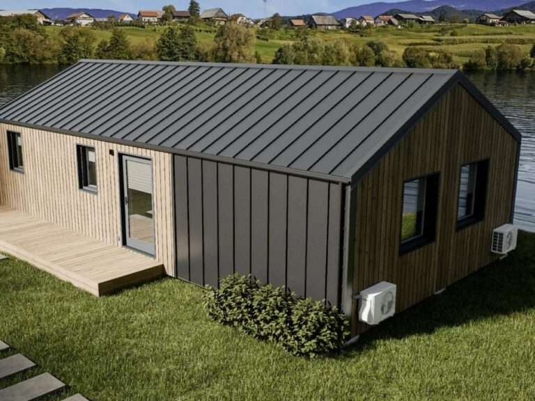 An elevated exterior view of the Vagabond Haven Evergreen XL prefab home, showcasing its natural wood cladding, dark metal standing seam roof, and large glass windows, set on green grass near a lake with a village in the background.