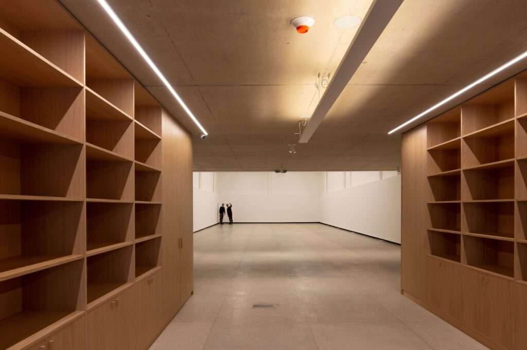 Symmetric indoor view of the Larivière Foundation library corridor lined with custom-built light oak wooden bookshelves, leading into the bright, open main exhibition hall.
