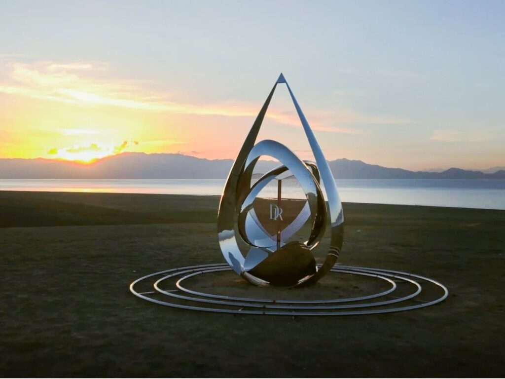 A teardrop-shaped reflective metal pavilion with interlocking rings at the center, situated on a grassy plain by Sayram Lake during a vibrant golden sunset.