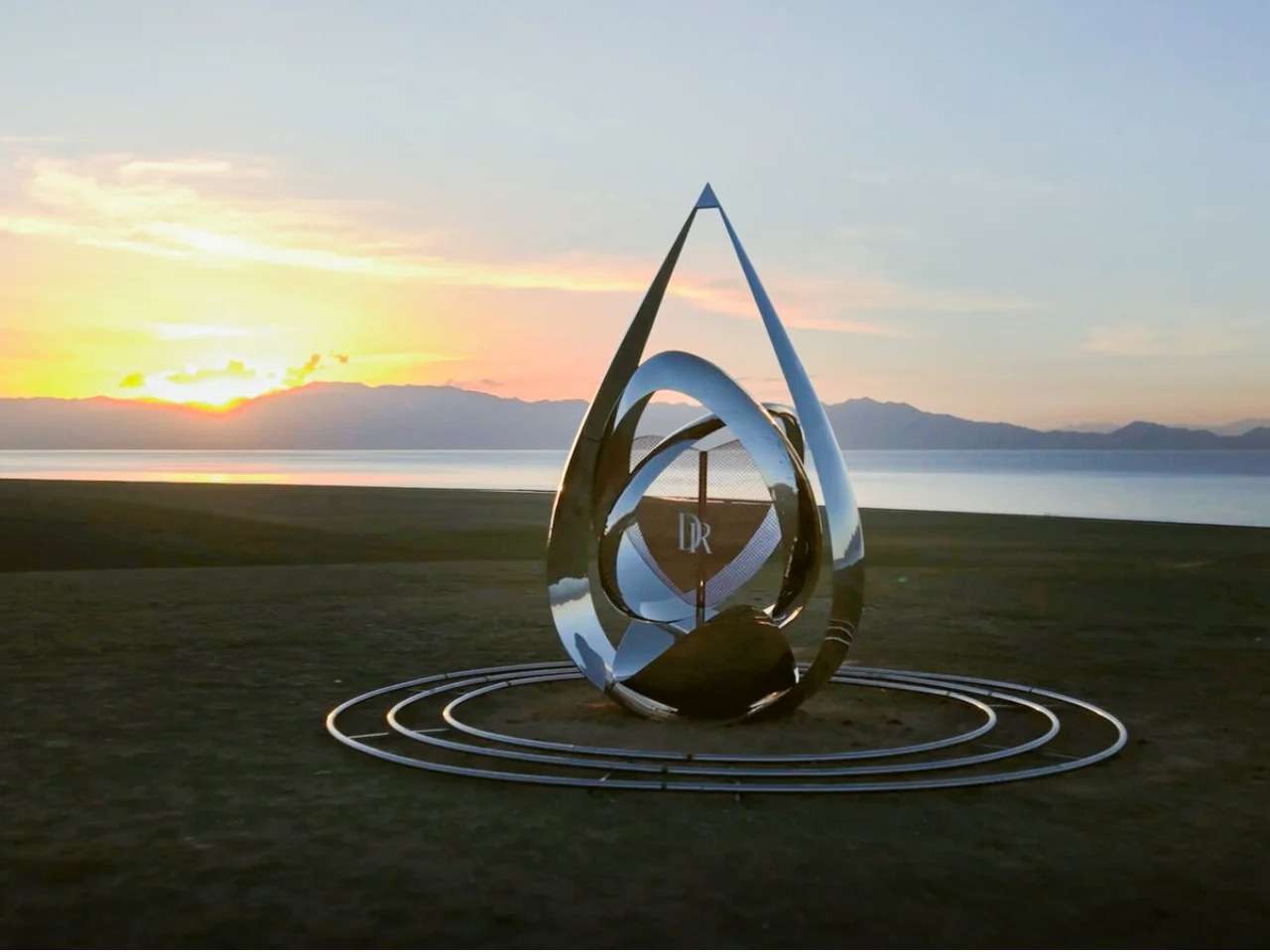A teardrop-shaped reflective metal pavilion with interlocking rings at the center, situated on a grassy plain by Sayram Lake during a vibrant golden sunset.