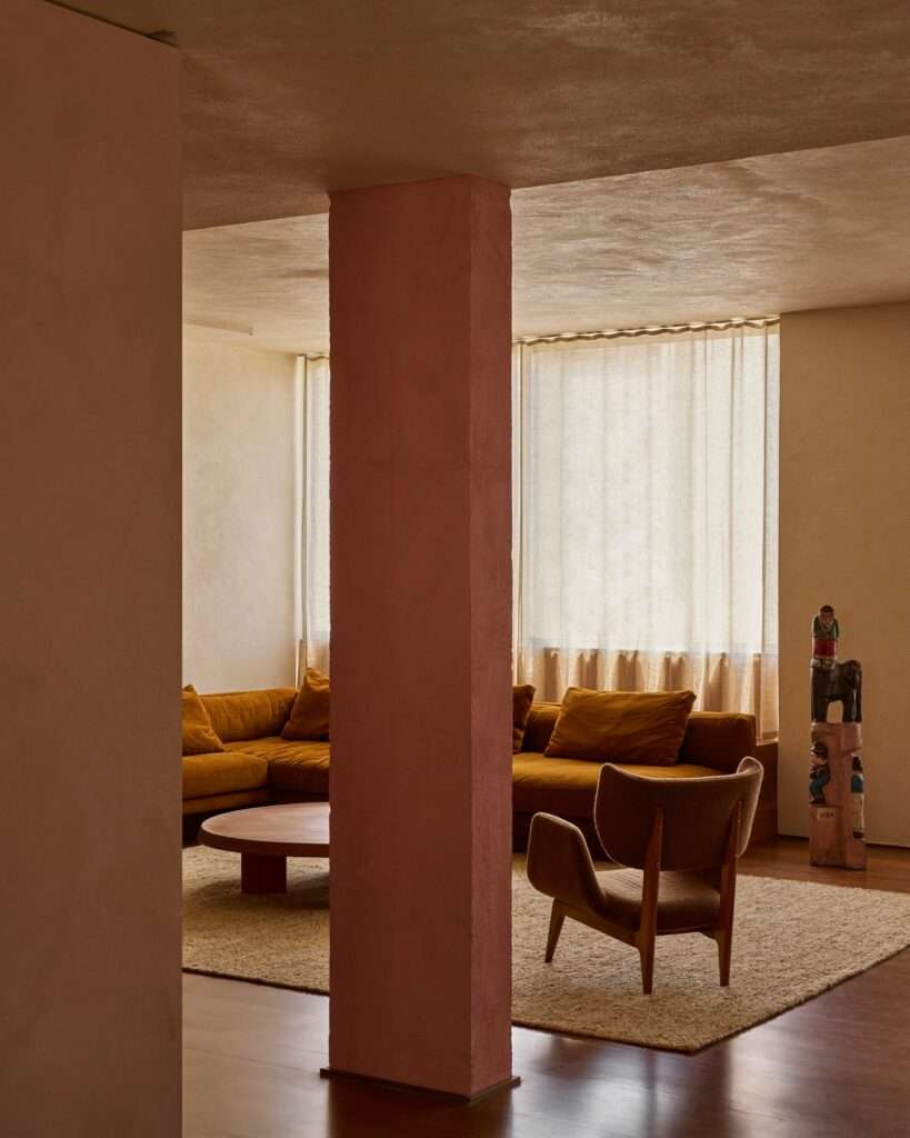 Close-up view of a prominent structural column painted in an intense pink color in an apartment living room, with a mustard velvet sofa, wooden coffee table, and an armchair in the background.