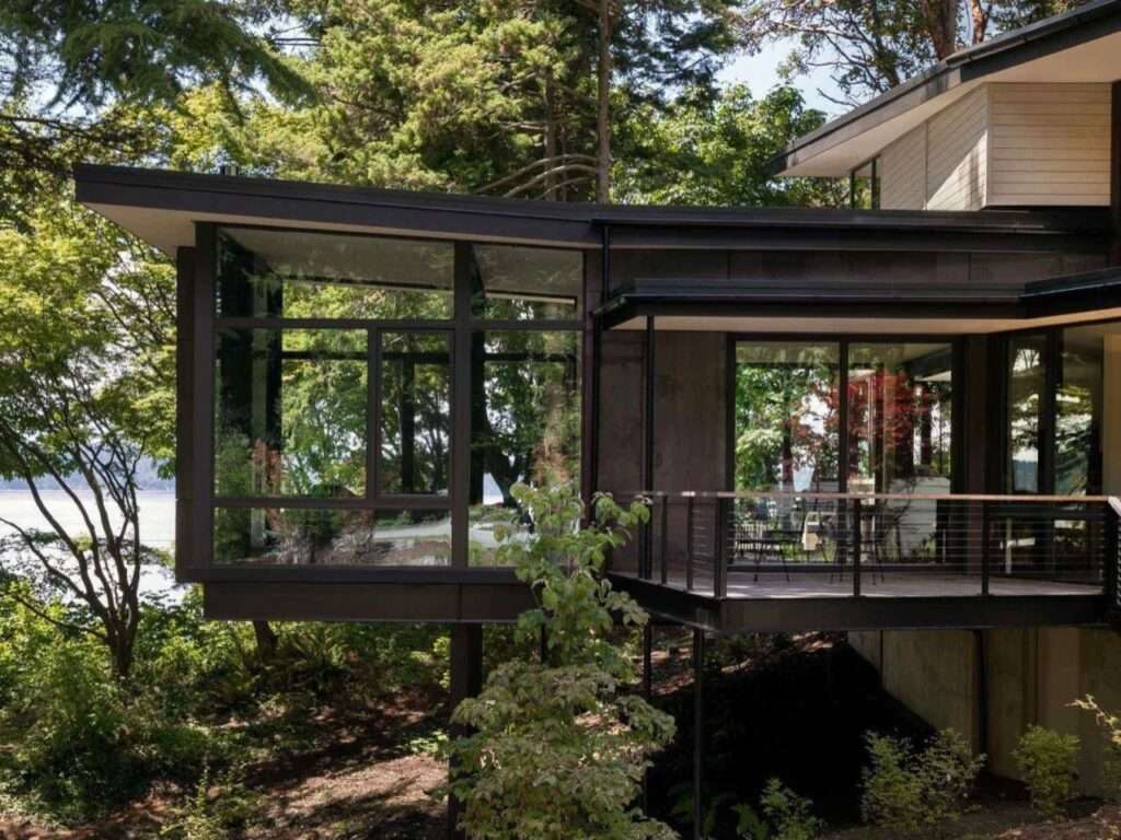 Modern cantilevered steel and glass residential volume extending over a forested slope with an attached outdoor deck.