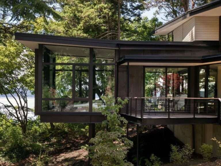 Modern cantilevered steel and glass residential volume extending over a forested slope with an attached outdoor deck.