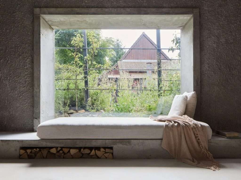 Built-in concrete window seat with neutral daybed cushion, blanket, and firewood storage niche looking out into a green garden.