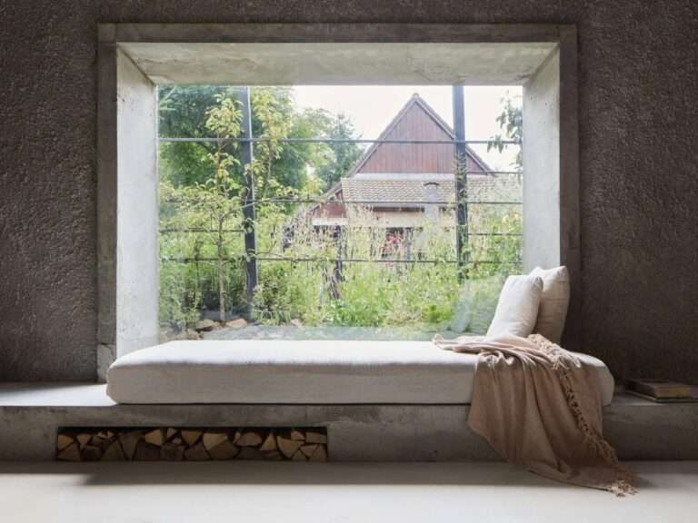 Built-in concrete window seat with neutral daybed cushion, blanket, and firewood storage niche looking out into a green garden.