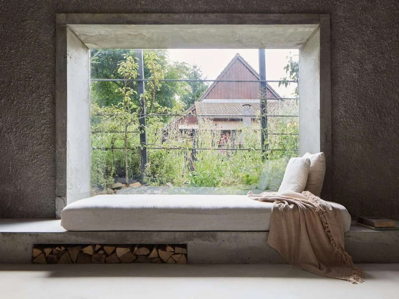 Built-in concrete window seat with neutral daybed cushion, blanket, and firewood storage niche looking out into a green garden.