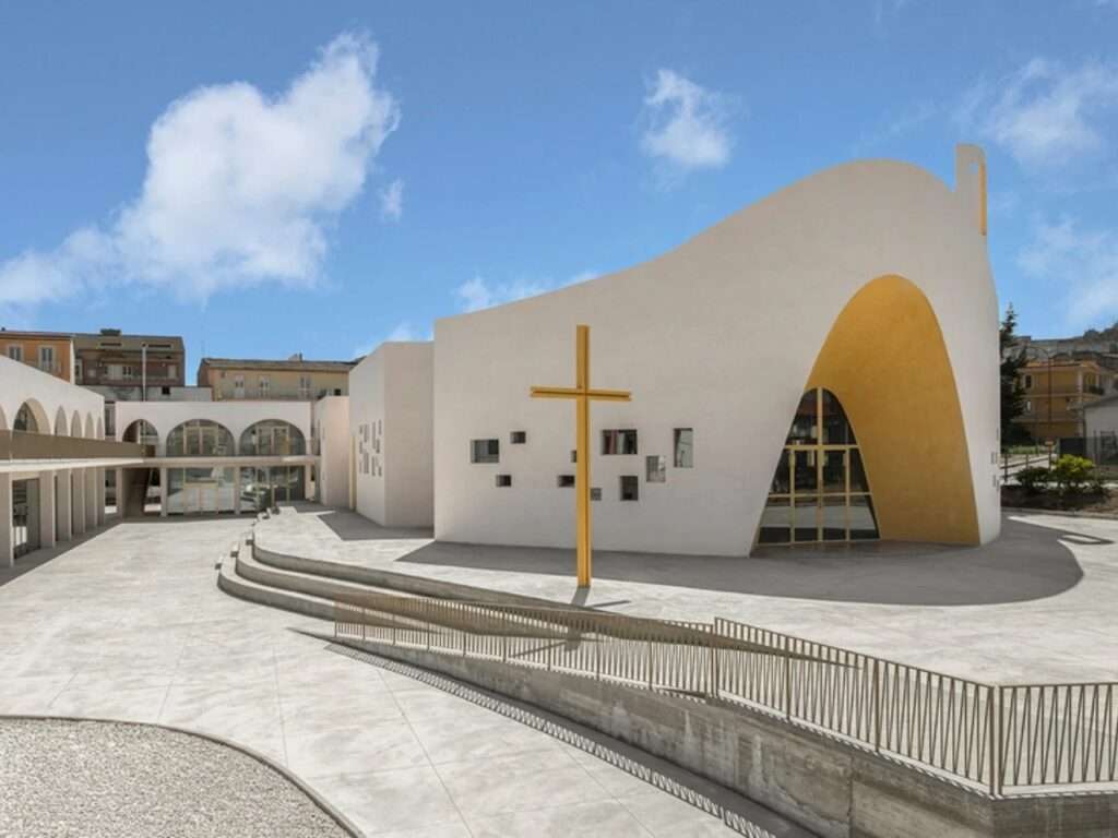 The main facade of the Santa Barbara Parish Complex in Licata with a large golden entrance arch, concrete courtyard, and a prominent freestanding golden cross.