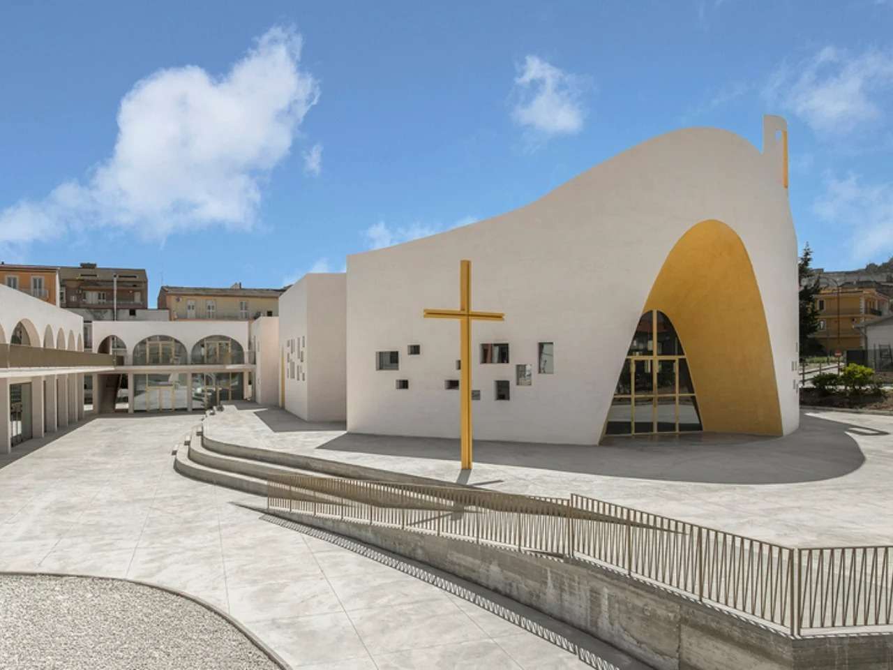 The main facade of the Santa Barbara Parish Complex in Licata with a large golden entrance arch, concrete courtyard, and a prominent freestanding golden cross.