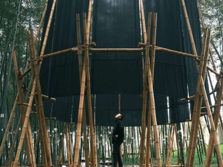 A visitor standing inside the temporary Veil Tower bamboo pavilion, looking up at the open vertical structure surrounded by dark textile membranes and a Moso bamboo forest.