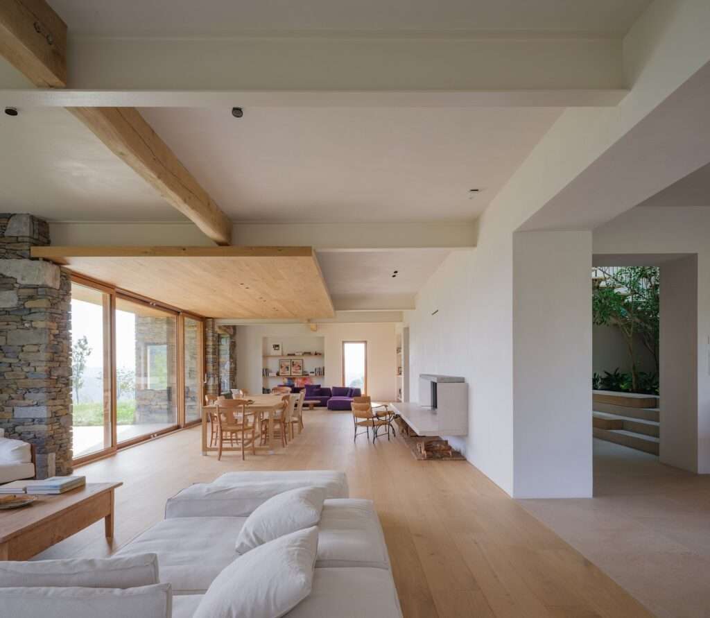 Modern open plan living and dining room with oak wood flooring, a minimalist white fireplace, and a partial view of an indoor courtyard with green plants.