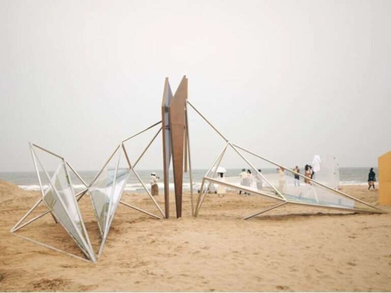 A lightweight winged pavilion structure made of interconnected metal rods and acrylic lightboxes on a sandy beach under a cloudy sky.