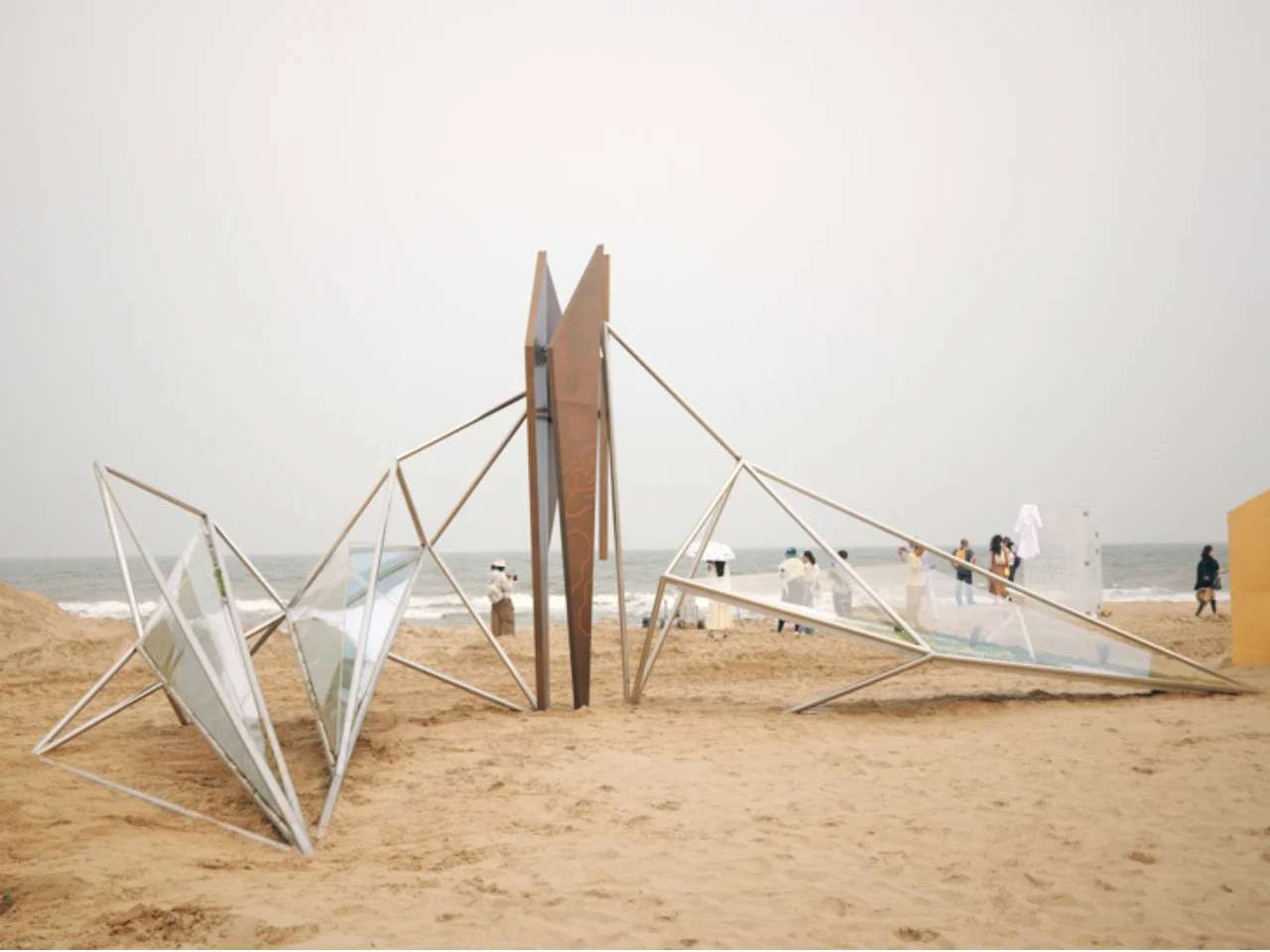 A lightweight winged pavilion structure made of interconnected metal rods and acrylic lightboxes on a sandy beach under a cloudy sky.