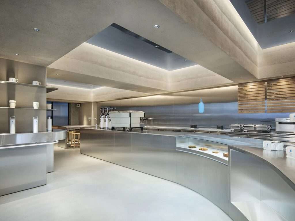 Minimalist espresso preparation area with stainless steel surfaces, illuminated blue bottle logo, and warm textured ceiling panels at Blue Bottle Coffee Harajuku.