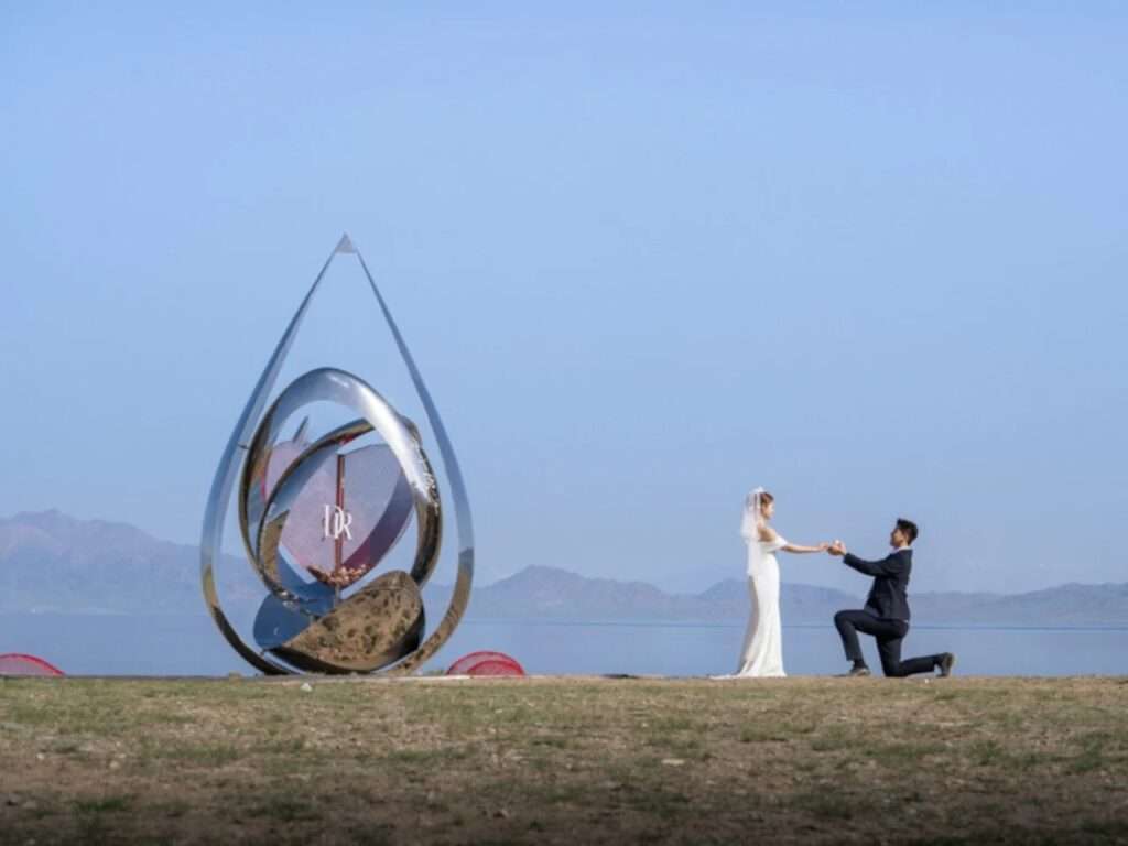 A man kneeling to propose to a woman in a white wedding dress next to the chrome water-droplet architectural installation at Sayram Lake under a clear blue sky.