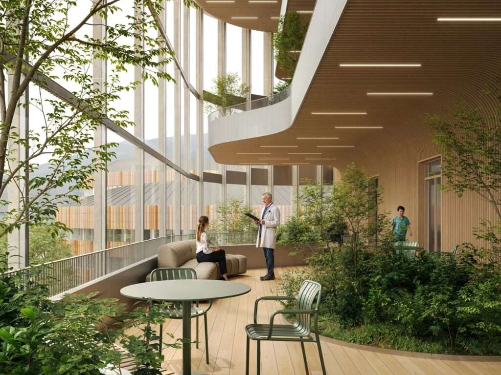 Doctor speaking to a patient on an indoor balcony filled with lush green plants, warm wood ceiling curves, and floor-to-ceiling windows.