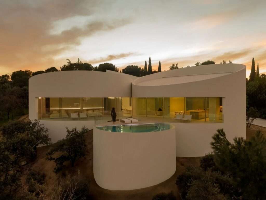 Exterior dusk view of House in Las Rozas featuring curved white cylindrical volumes, panoramic glass windows, and an elevated circular swimming pool overlooking the landscape.