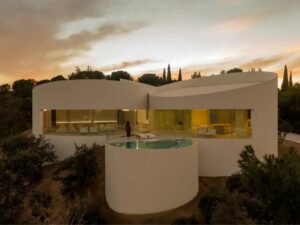 Exterior dusk view of House in Las Rozas featuring curved white cylindrical volumes, panoramic glass windows, and an elevated circular swimming pool overlooking the landscape.