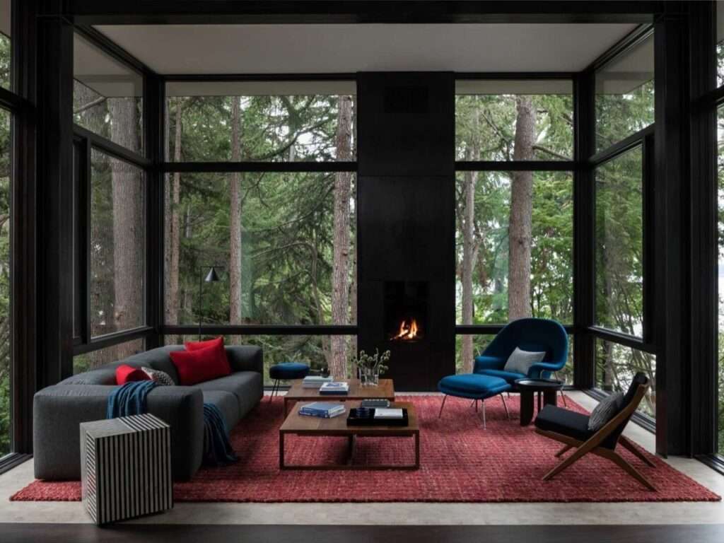 Suspended modern living room featuring floor-to-ceiling steel-framed windows, a black fireplace, a red rug, and a blue accent chair facing a dense forest.