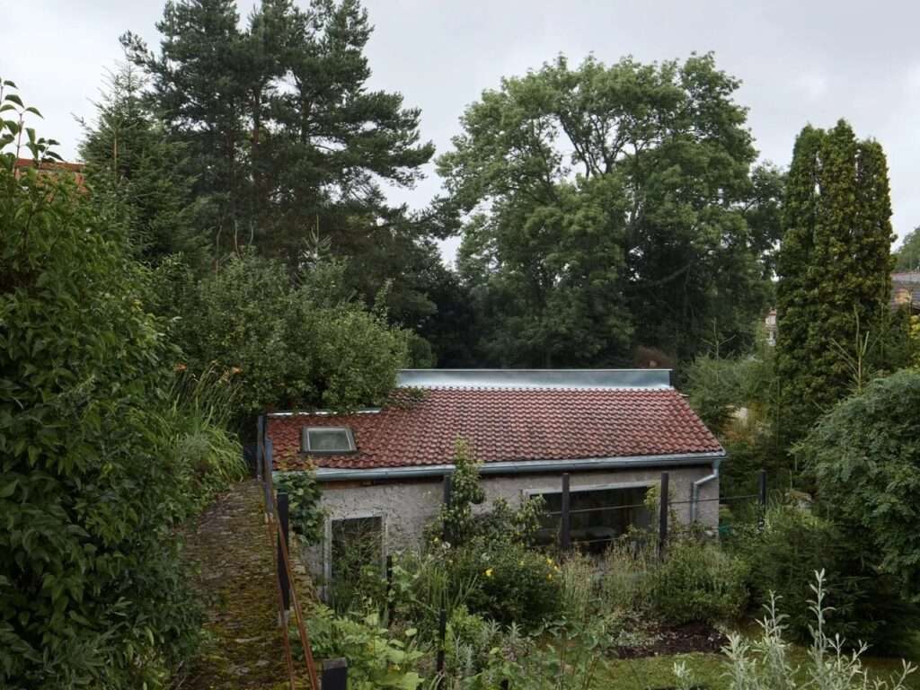 High-angle exterior view of a small repurposed stone summer house with red tile roof surrounded by dense trees and plants.