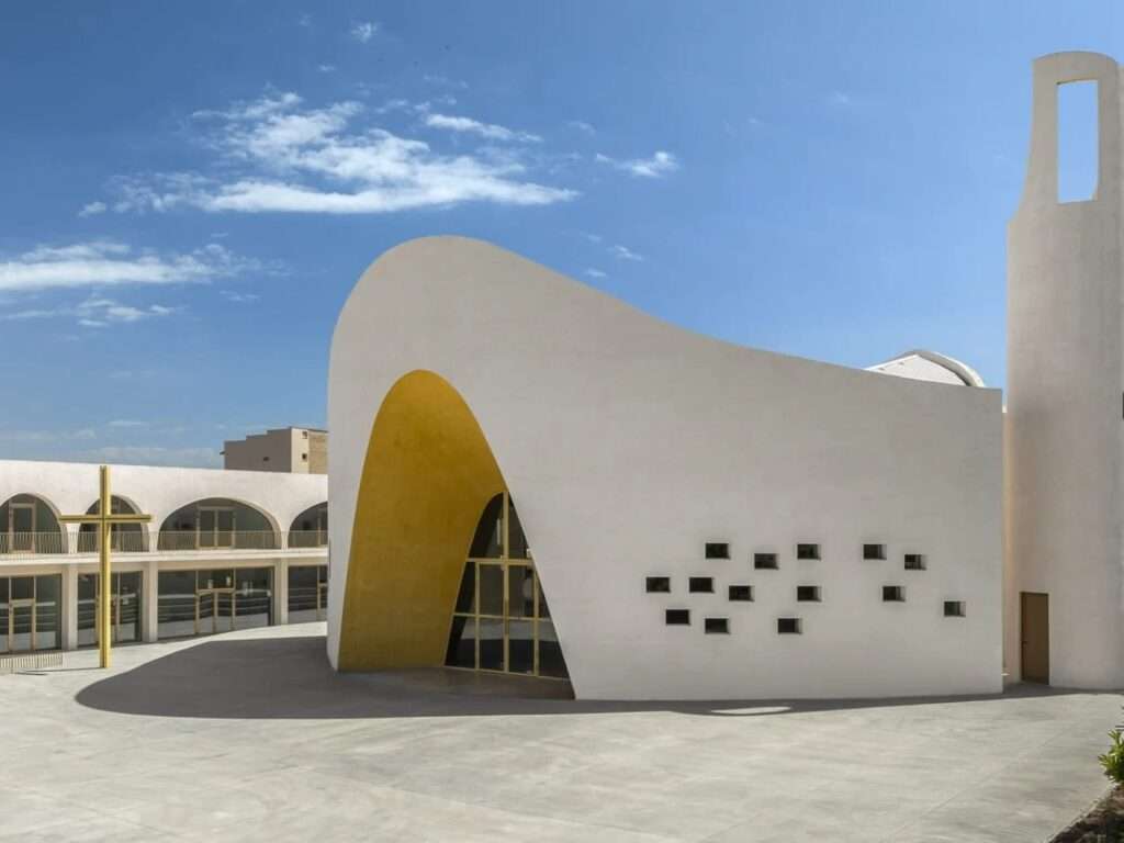 Side perspective of the white curved concrete church building of Santa Barbara Parish with small square window openings and an integrated bell tower.