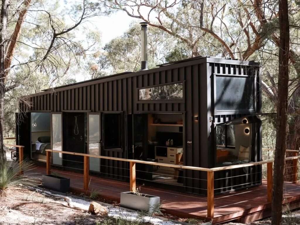 Dark vertical clad off-grid cabin SHAK in the Australian Grampians bush, showing its angled roofline, surrounding wooden deck with railing, planters, and open doors revealing the interior.