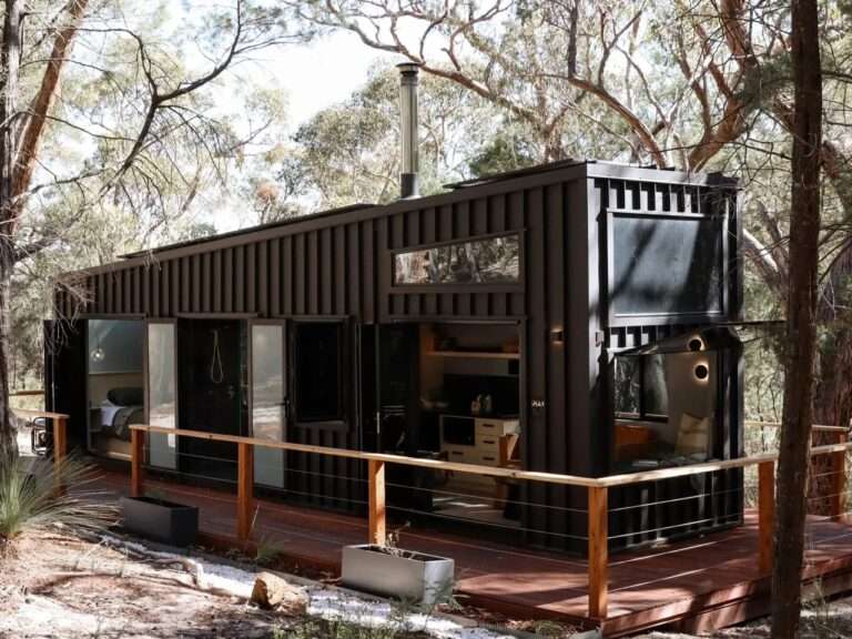 Dark vertical clad off-grid cabin SHAK in the Australian Grampians bush, showing its angled roofline, surrounding wooden deck with railing, planters, and open doors revealing the interior.
