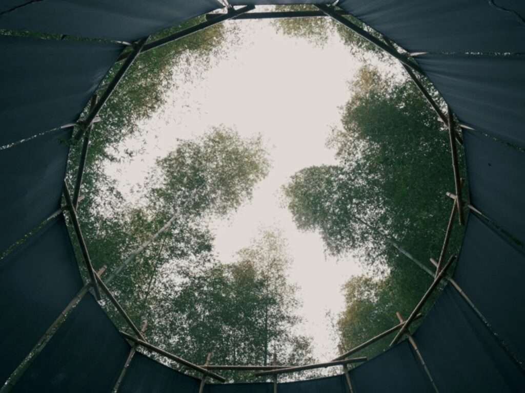 Low-angle view looking up through the circular open roof crown of the bamboo structure framed by dark textile panels against the sky and Moso bamboo treetops.