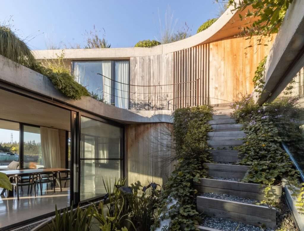 An architectural interior viewpoint looking through a floor-to-ceiling curved glass curtain wall toward a private central courtyard with rustic wooden stairs and lush vegetation.