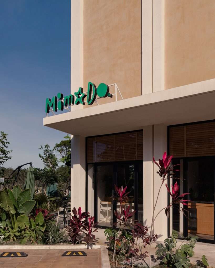 Modern exterior facade of Mimado Restaurant featuring green neon typography signage, tall glass entry door, and tropical landscaping.