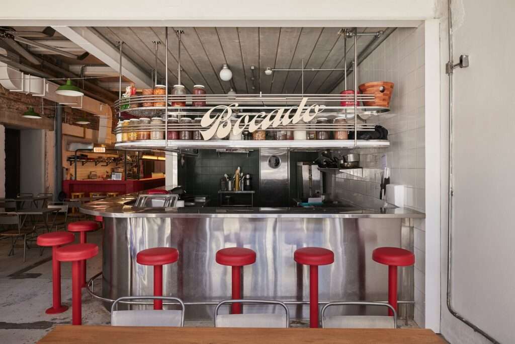 Adaptive reuse of Fábrica Bhering in Rio de Janeiro, Brazil. Close-up view of the "Bocado" restaurant's central bar and kitchen area. The design features a stainless steel counter, overhead storage for preserved foods, and distinctive red bar stools. Photographed by Rafael Salim.