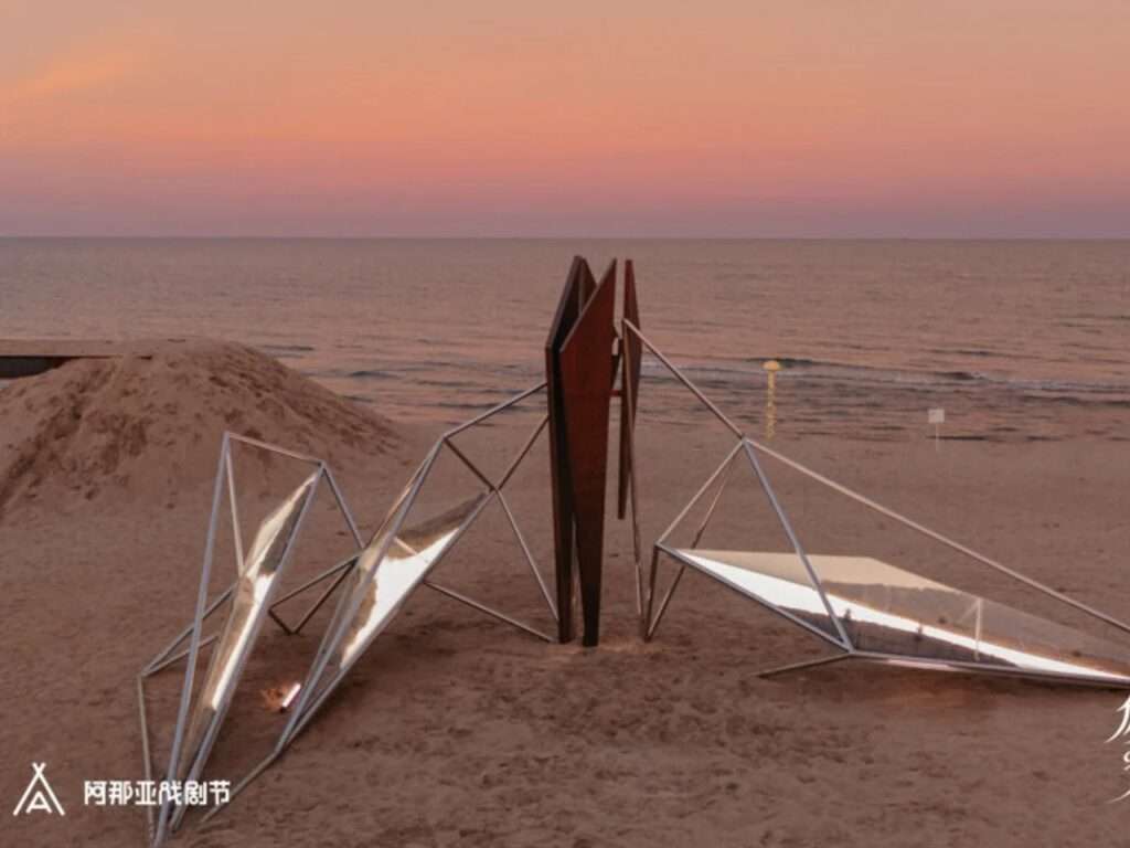 Architectural pavilion structure on a beach shoreline at sunset with reflective acrylic panels mirroring the orange sky.