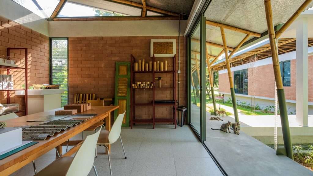 Internal workspace within the studio at Anjung House, highlighting a raw wooden conference table, exposed compressed earth blocks, and a large sliding glass door looking out toward the garden.