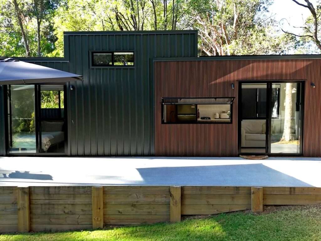 Exterior view of the Burley 9.6 tiny home showcasing a mix of dark vertical corrugated metal siding and warm NewTechWood slatted timber cladding next to a concrete patio.