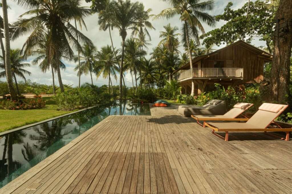 Outdoor wooden pool deck with sun loungers beside a long lap pool surrounded by coconut palms.