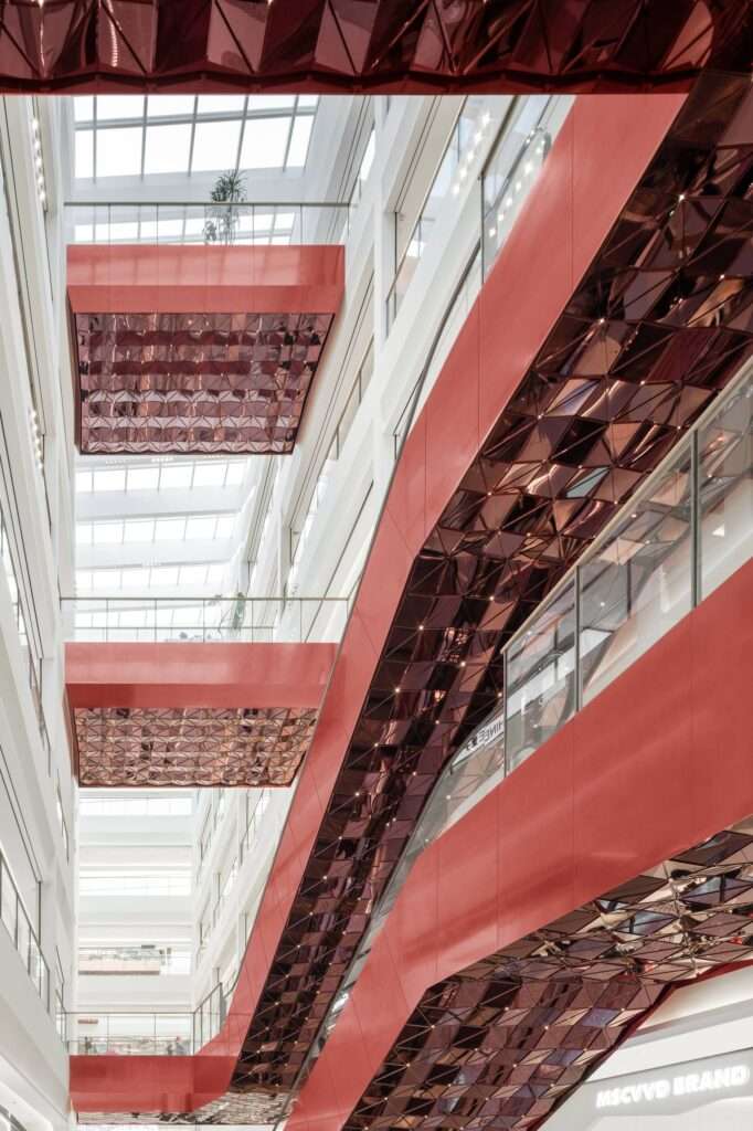 Low-angle close-up view of sculptural red escalators and bridge structures under a bright skylight at Wuxi MixC Mall, showing faceted reflective metallic under-claddings.