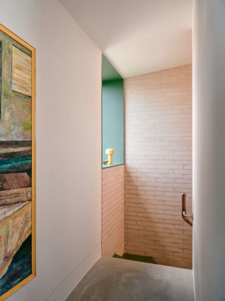 Light-colored brick stairwell with a curved wall, wooden handrail, and a small green recessed window nook holding a yellow lamp.