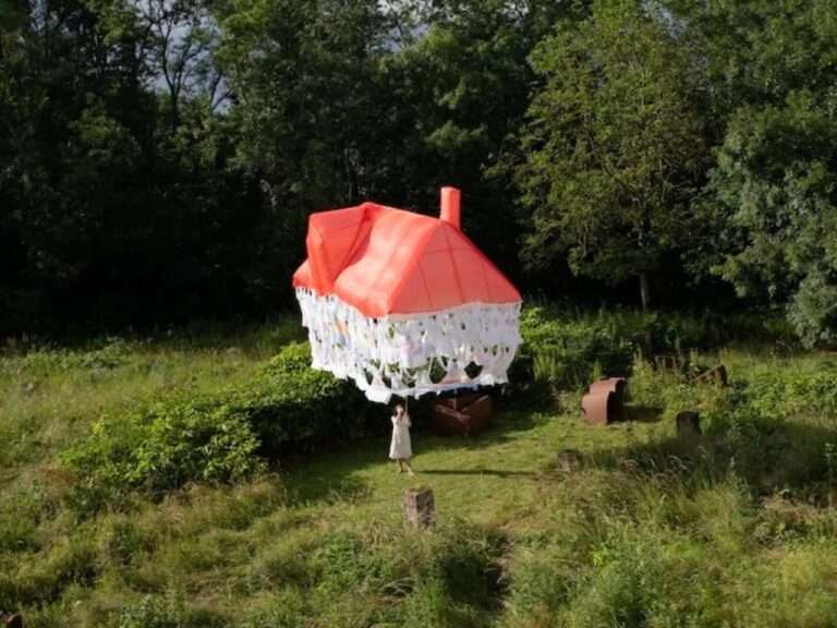 A woman in a white dress standing in a green field, holding a thin rope attached to a floating, lightweight installation shaped like a house with a red translucent roof and a skirt made of interconnected white discarded clothes.
