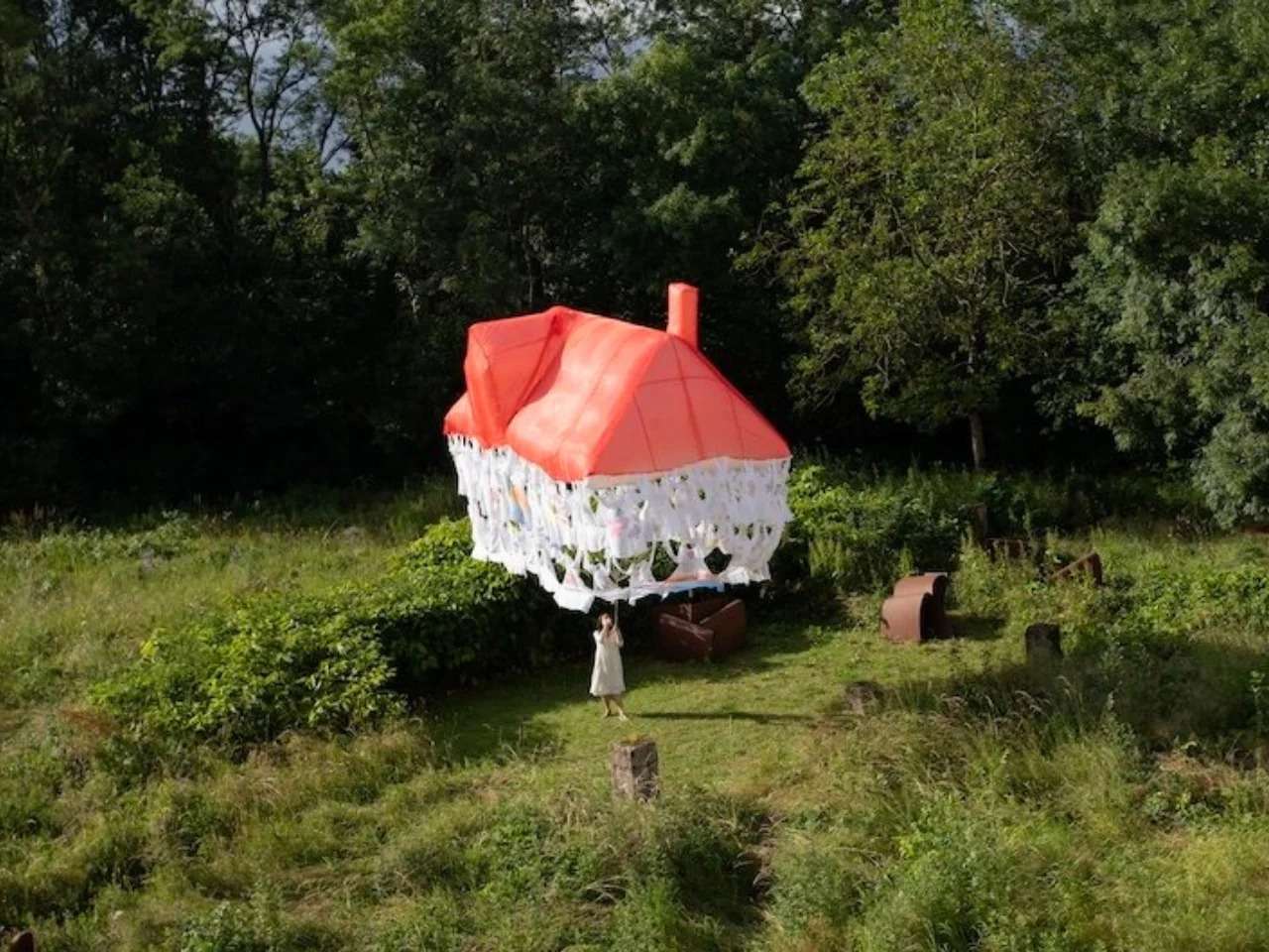 A woman in a white dress standing in a green field, holding a thin rope attached to a floating, lightweight installation shaped like a house with a red translucent roof and a skirt made of interconnected white discarded clothes.